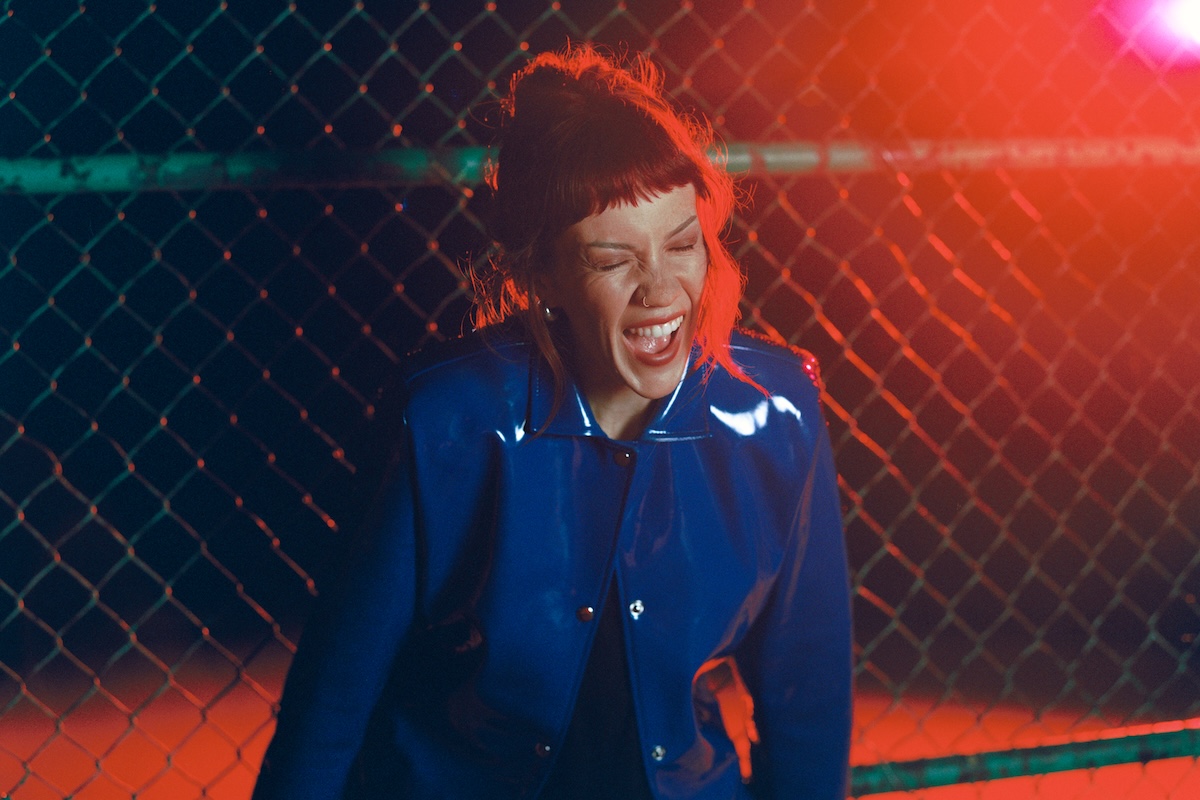 Singer-songwriter Bess Atwell smiling in a blue patent jacket against a chain-link fence under red lighting, photo by Elizabeth Landry.